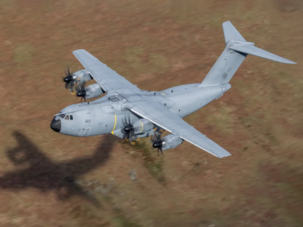 A large, grey military cargo plane with yellow strips flying close to rural ground.