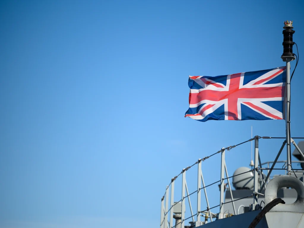 A close-up of a Union Jack waving in the wind while fixed to the bow of a large military vessel.