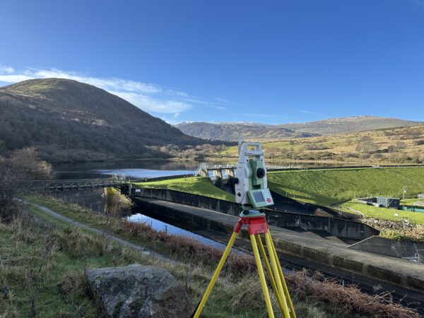 A geospatial total station positioned on a grassy hillside with a bridge in the background, stretching over a water source.