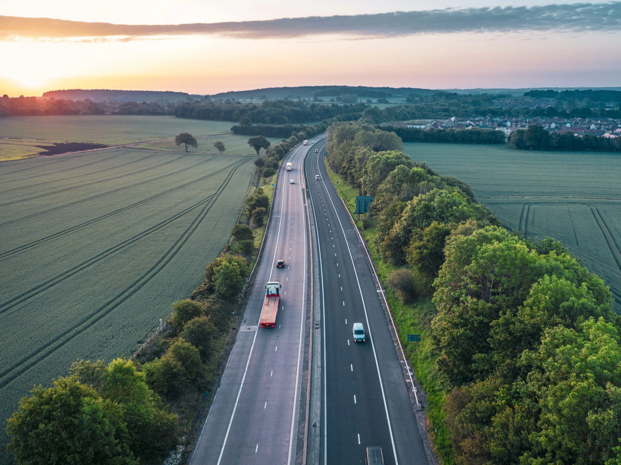 A quiet motorway between two large, green fields and lined with trees either side.