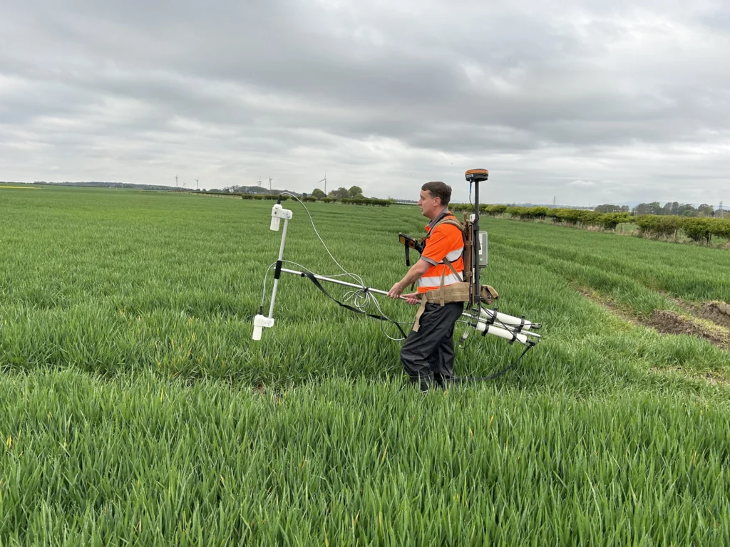 A land surveyor carrying equipment through a large, grassy field on a cloudy day.