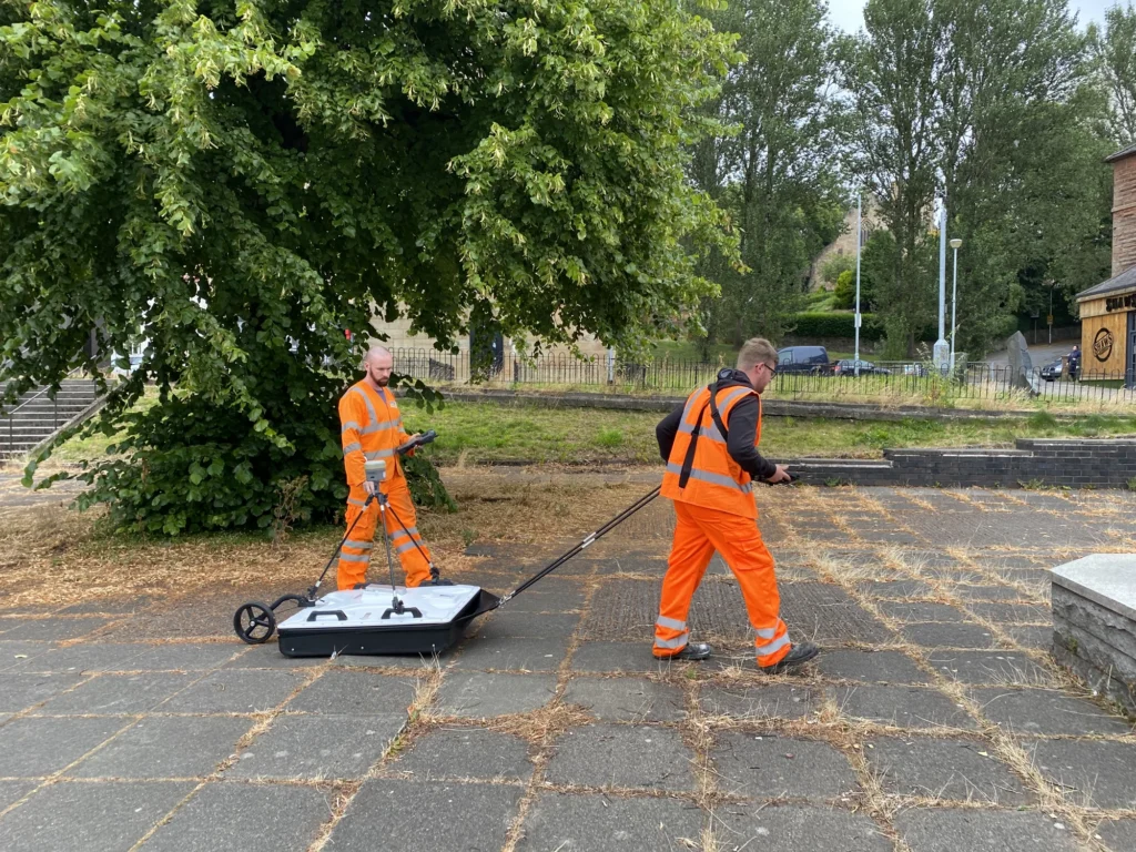 Two land surveyors in orange high-vis gear, walking a piece of equipment across a concrete slabbed area.