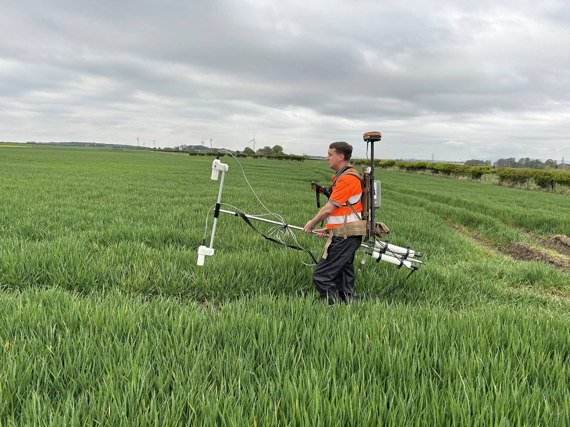 A land surveyor carrying equipment through a large, grassy field on a cloudy day.